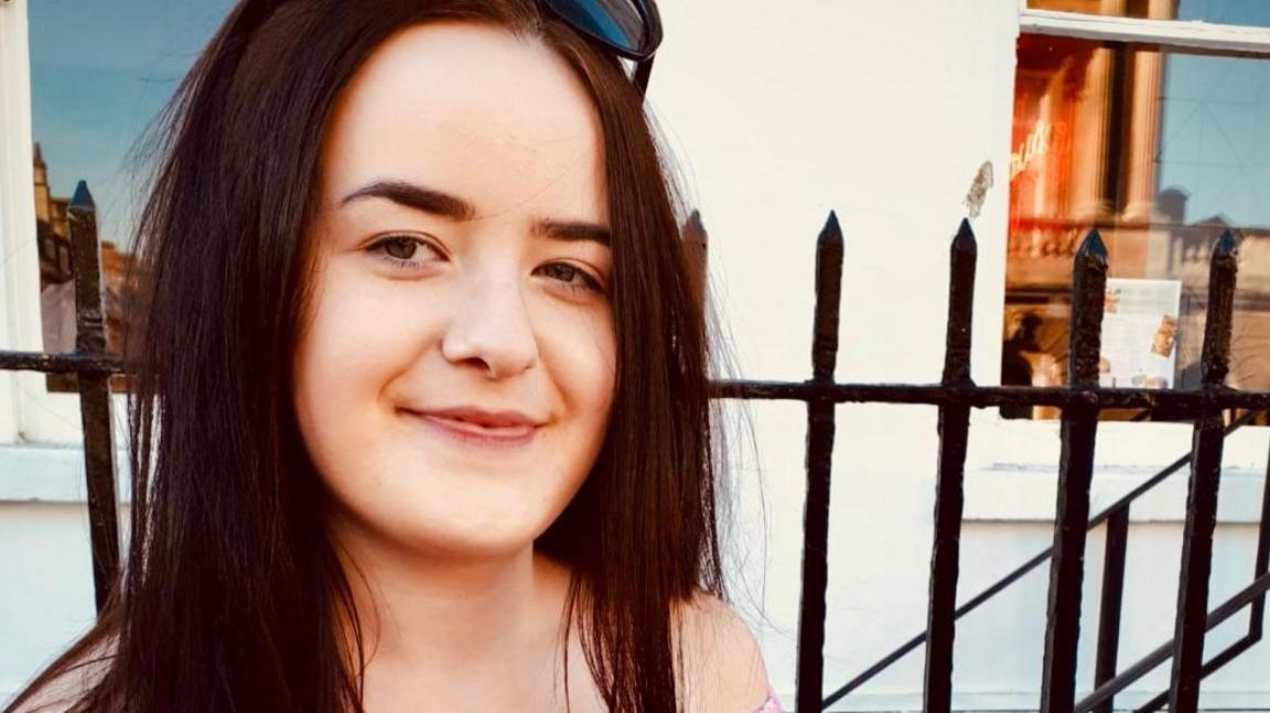 Emily Griffiths is sitting outside, with black railings and a white buiding in the background. She has long dark hair and is smiling at the camera.