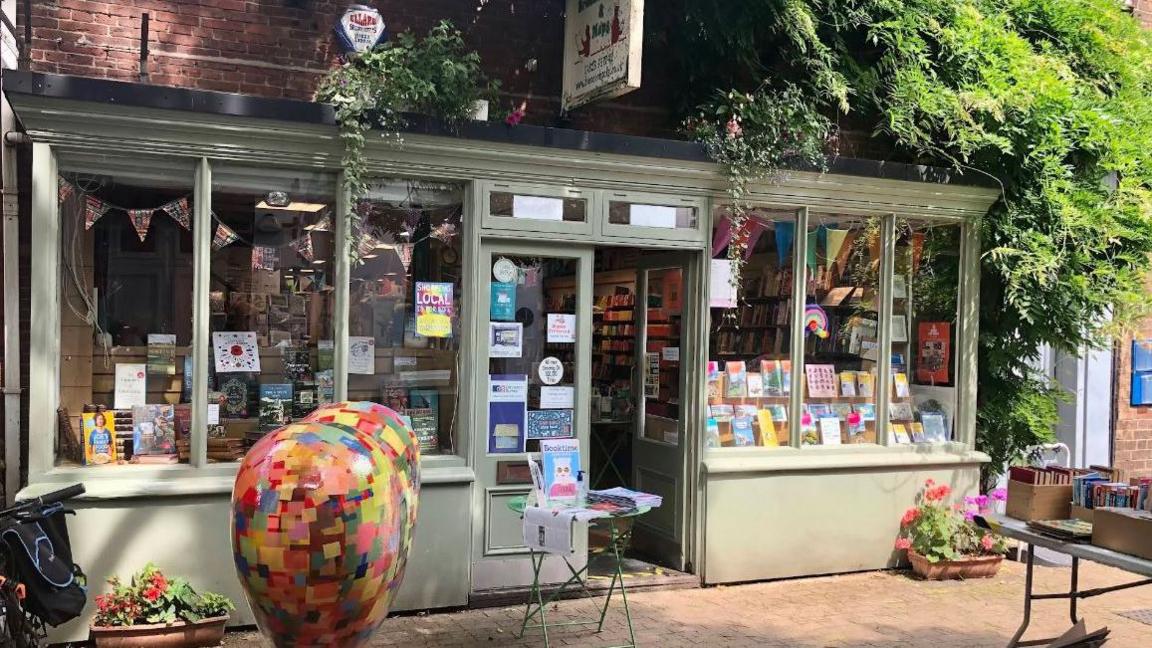 The exterior of the Brendan Books shop in Taunton. It is a quaint green shop with large windows at the front and double doors. It is tucked away in a sunny brick courtyard beneath thick fern and ivy plants growing over the doorway. There are books on display in the windows, bunting hanging above, and flower planters outside. 