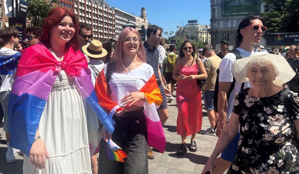 Young women at Pride sporting colourful banners and an elderly lady in a floral dress and sunhat