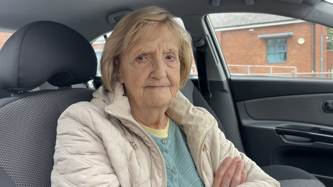 A close up shot of an elderly woman wearing a beige coat with a blue jumper underneath. She has her arms crossed. She has short blond hair. She is sat in her car.