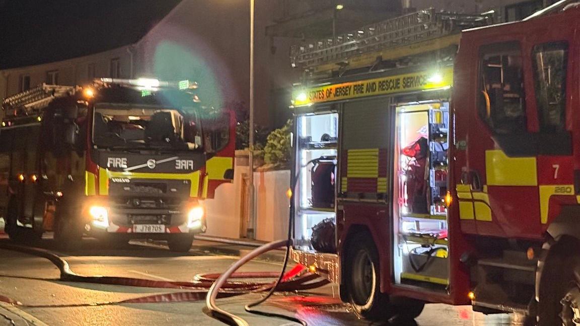 Two fire engines are stationary on the side of a road with hoses coming out from them and laying across the road. It is dark and a row of houses can be seen behind them.