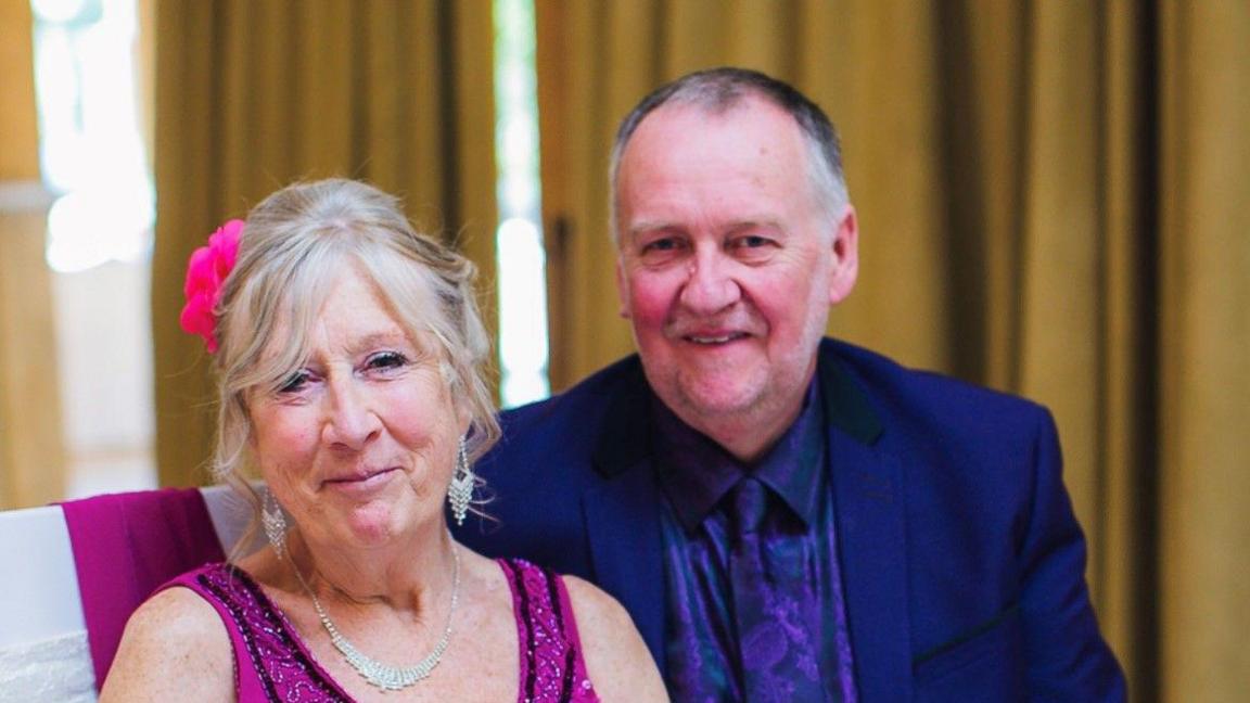 Thelma Huse wearing a pink sparkly dress and a pink flower in her hair. She is smiling and sitting beside Tony Mist, who is wearing a dark blue suit and looking at the camera