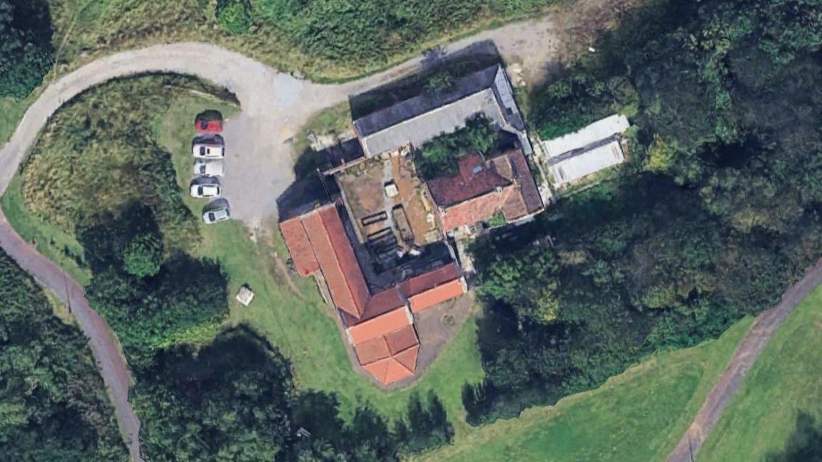 An aerial view of a farmhouse, surrounded by fields and cars on a driveway.