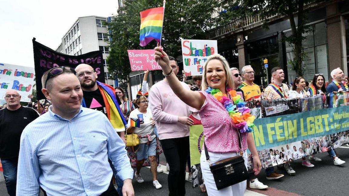 Michelle O'Neill is in the centre of the image taking part in 2022 Belfast Pride. She is wearing a pink top and a rainbow flower garland and white trousers and holding the rainbow flag. Around her are other marchers including a group of people holding a banner which says "Sinn Féin LGBT" in light green letters on a darker green background.