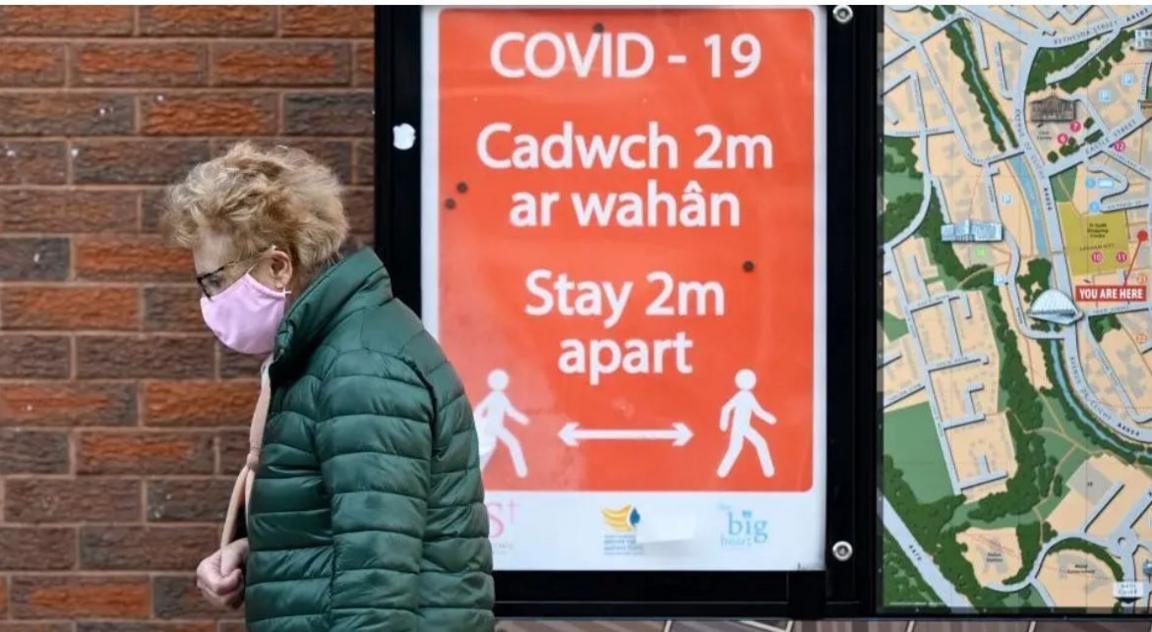 A woman in a face mask walking past a sign during the Covid pandemic asking people to stay two metres apart 