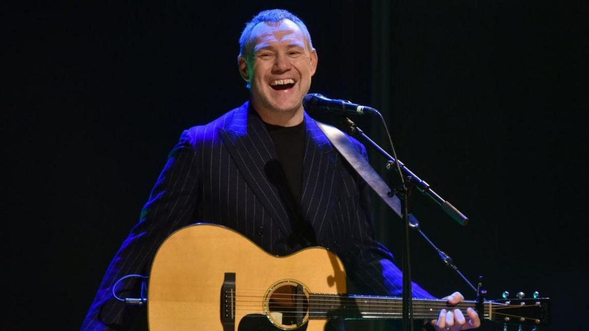 David Gray performing on a stage. He is in a dark pin strip suit holding a brown acoustic guitar. There is a dark blue light on him. 