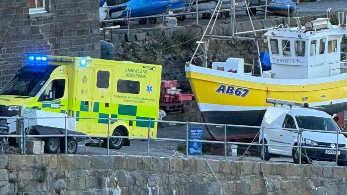 A pier with a docked yellow and balck boat, and a yellow ambulance parked up next to it.