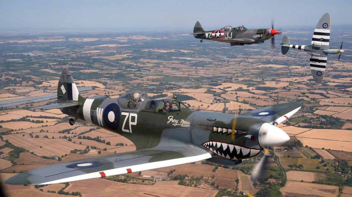 Three Spitfires fly in formation above the Kent countryside. The aircraft in the foreground bears shark-mouth nose art and the name "Grey Nurse", while two other Spitfires fly behind it over a patchwork landscape of fields, roads and villages.