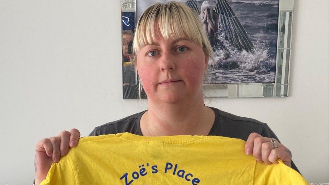 Stephanie Perry, with blonde hair cut into a fringe, looks at the camera with a serious expression and holds up a yellow t-shirt with the Zoe's Place logo