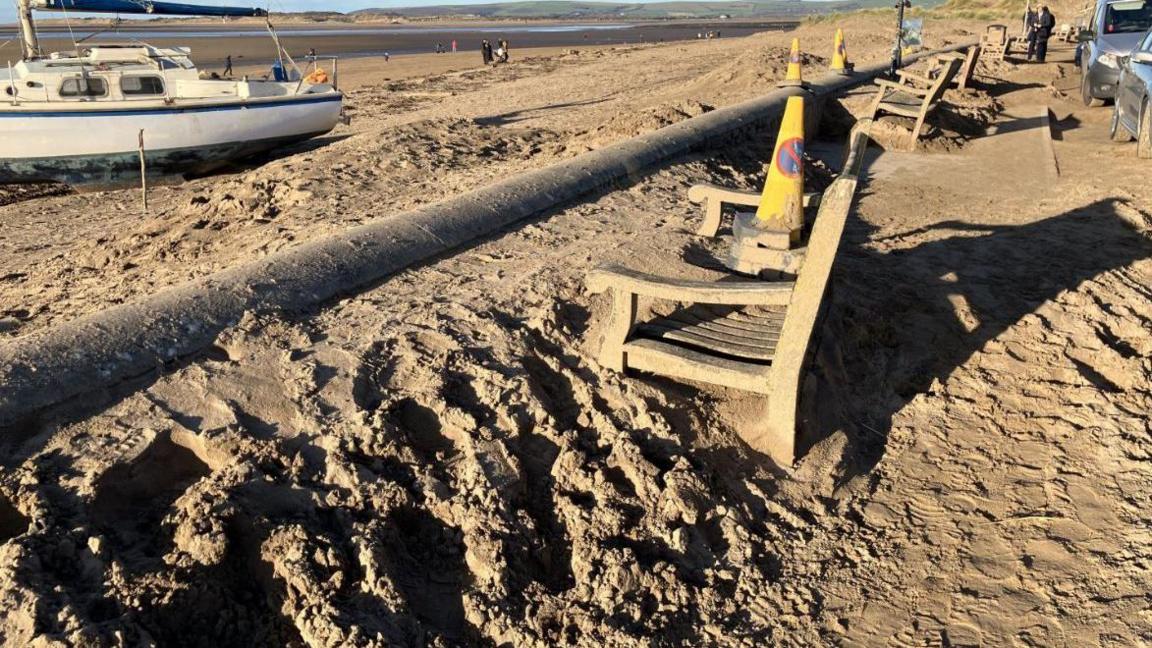 A large amount of sand has covered a pavement with a set of four benches on it, which are difficult to sit on due to the sand. Yellow cones have been put on the benches. A white yacht is moored up on the opposite side of a wall by the benches.