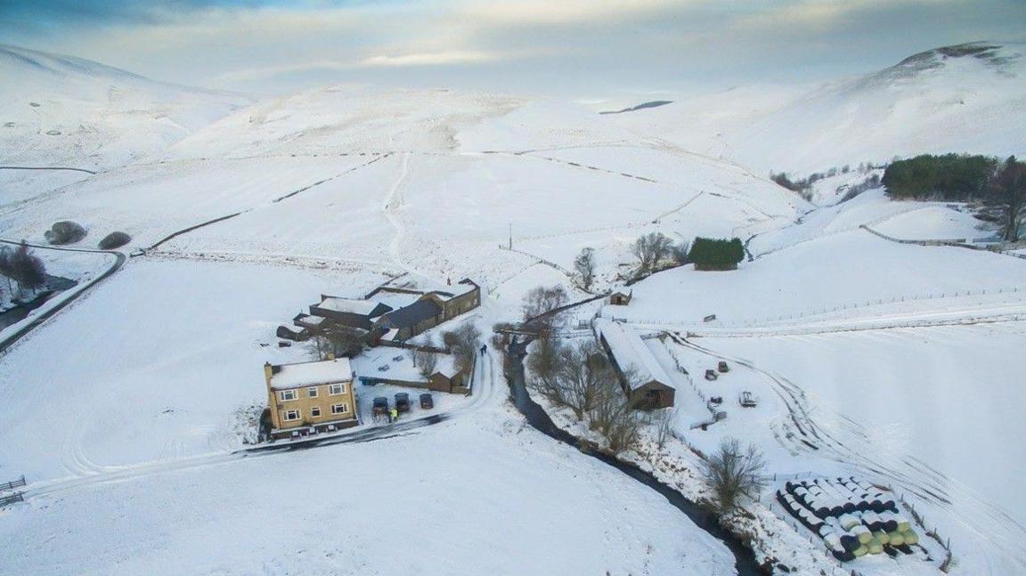 A drone image of a remote farm surrounded by mountains covered in snow with some bales of straw visible to the right 