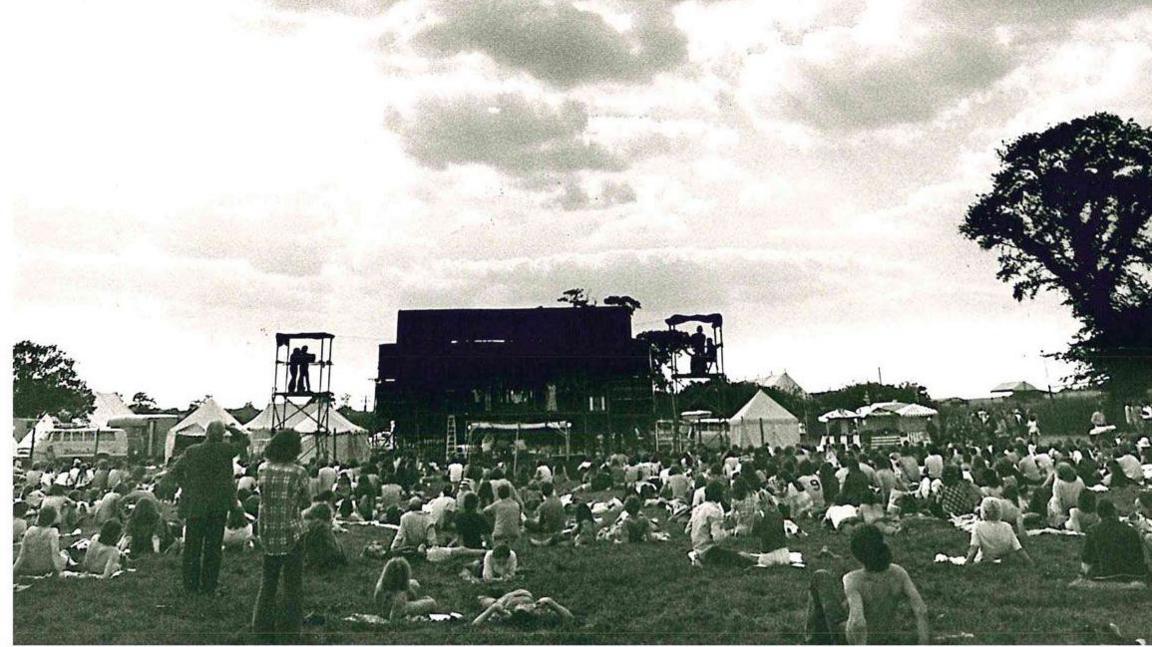 A black and white photo with a dark-coloured main stage in the centre. There is a scaffolding tower to the left supporting a camera and operator. There are small marquees either side of the stage. A crowd of people is sitting on the grass, watching the stage.