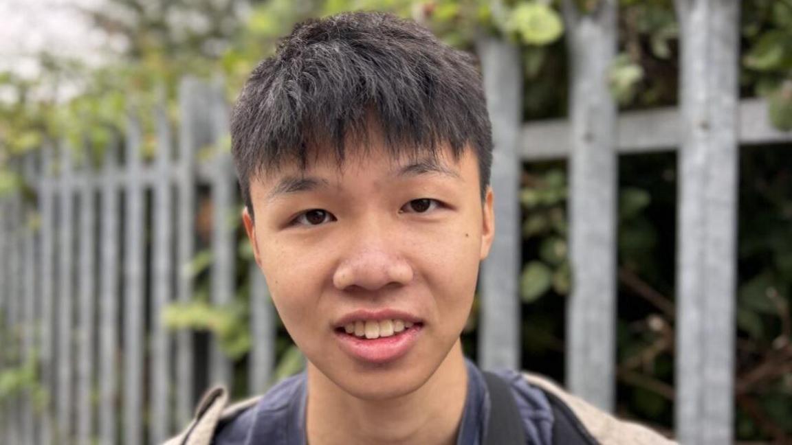 A man with short black hair with a fringe is smiling into the camera in a head shot. Behind him is a blurred background of a silver fence and trees. He is wearing a navy top and jumper.