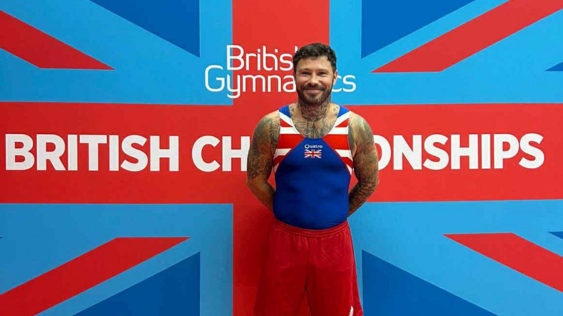 A man wearing a blue, red and white sports vest and shorts with the Union flag as part of the kit. He has short brown hair, a brown beard and tattoos on his arms. He is standing in front of a Union flag banner with "British Gymnastics British Championships" written on it in white.