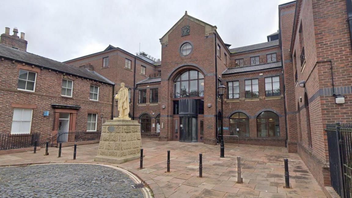 A general street view of the front of the Carlisle Crown Court red brick building. It has the royal coat of arms above the front door, there is a statue outside and the street is a mix of cobbled and paved.