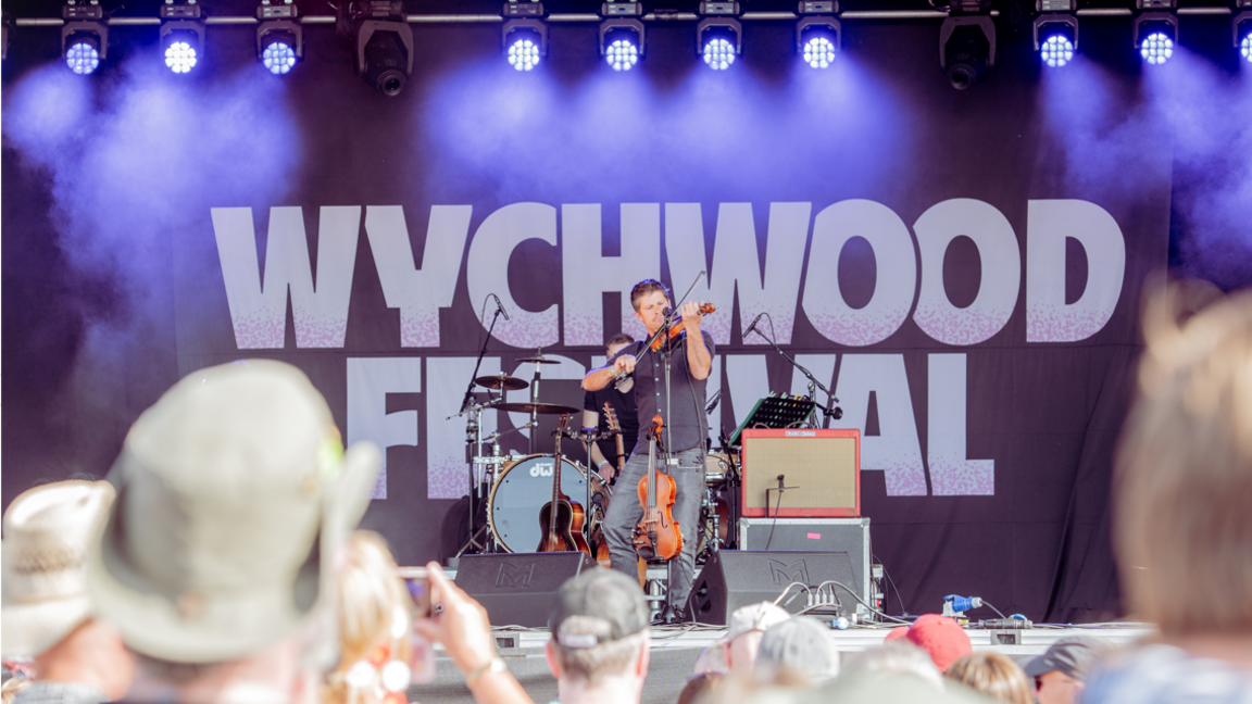 A violinist plays on a stage with Wychwood Festival written behind him and a crowd watching on a bright sunny day