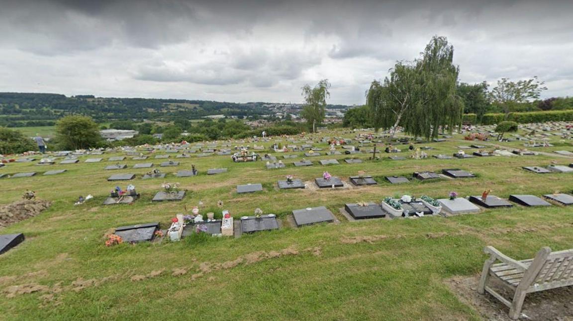 A view looking down the hill at South Bristol Cemetery, with hundreds of grave sites and headstones visible in the grass. There are trees planted at the cemetery, and it has a landscape view of fields and hedgerows.