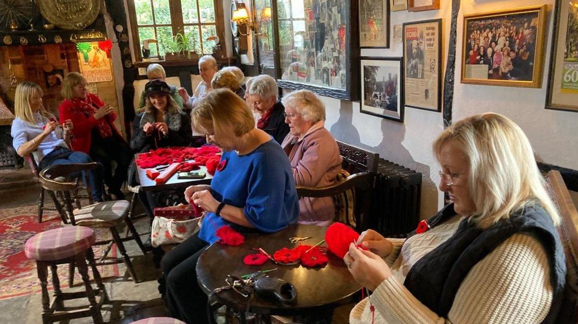 A group of women are knitting poppies in the foreground in a pub. Poppies are on the tables. There are framed photos on the walls and a rug. 