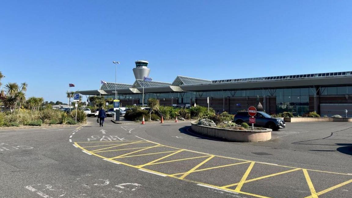 Jersey Airport on a sunny and clear blue sky day. There is a man walking up to the airport building which is largely glass with three triangular shaped glass roofs. The air traffic control tower, a silver spherical building, can be seen just past the airport.