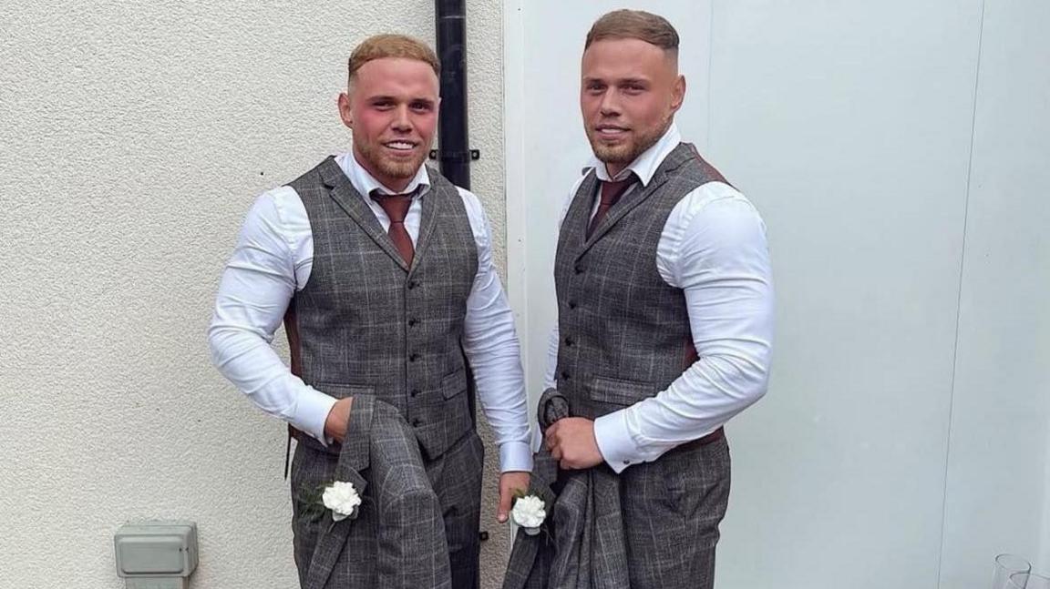 Corey and Liam dressed in white shirts and grey waistcoat and trousers with maroon tie. They both have short light brown hair and a short beard and are carrying grey jackets with white carnations in the buttonhole.