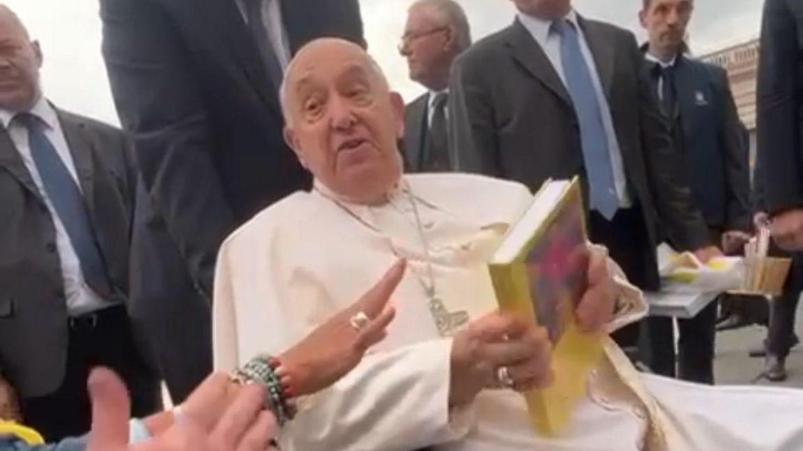 The pope looks into the camera talking to George White and holding a book given to him by George entitled "Trans life in the catholic church today".