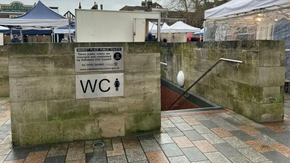 The underground toilet block. It shows a concrete wall and staircase leading below ground. It is positioned in the middle of a market square with stalls and marquees surrounding it. Two silver signs are on the concrete wall. The top ones says 'Market place public toilets' and explains they are managed by the council. The sign below says WC and has the symbol usually on women's toilets.