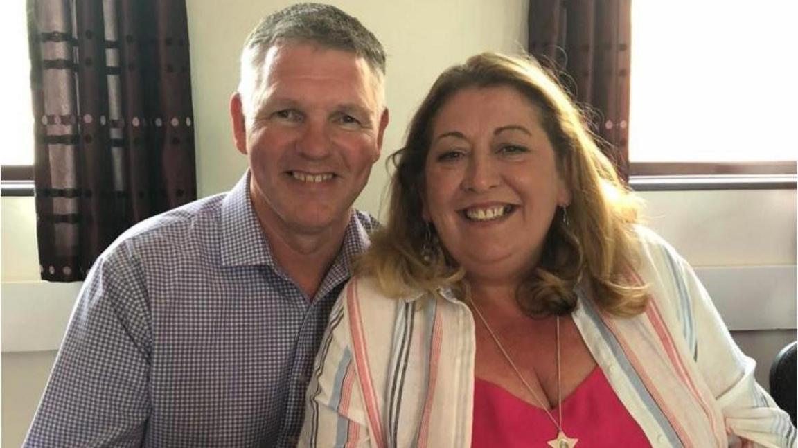 A man in a checked shirt smiling at the camera next to a woman also smiling at the camera. She is wearing a striped shirt with a red top underneath