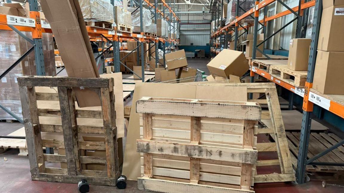 Several wooden pallets are propped up against each other in a warehouse next to an aisle of empty goods other than cardboard boxes.