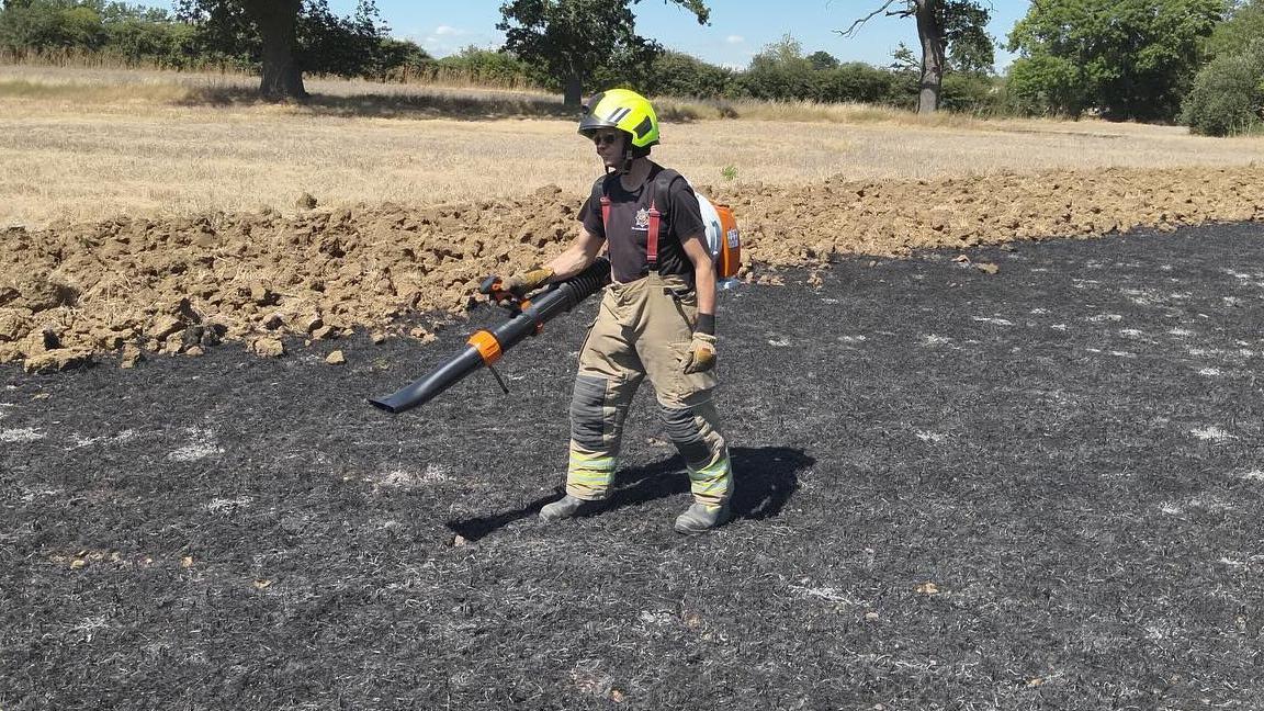 A firefighter, hold a piece of equipment, wearing protective trousers, gloves, a black T-shirt and helmet. They have equipment on their back.