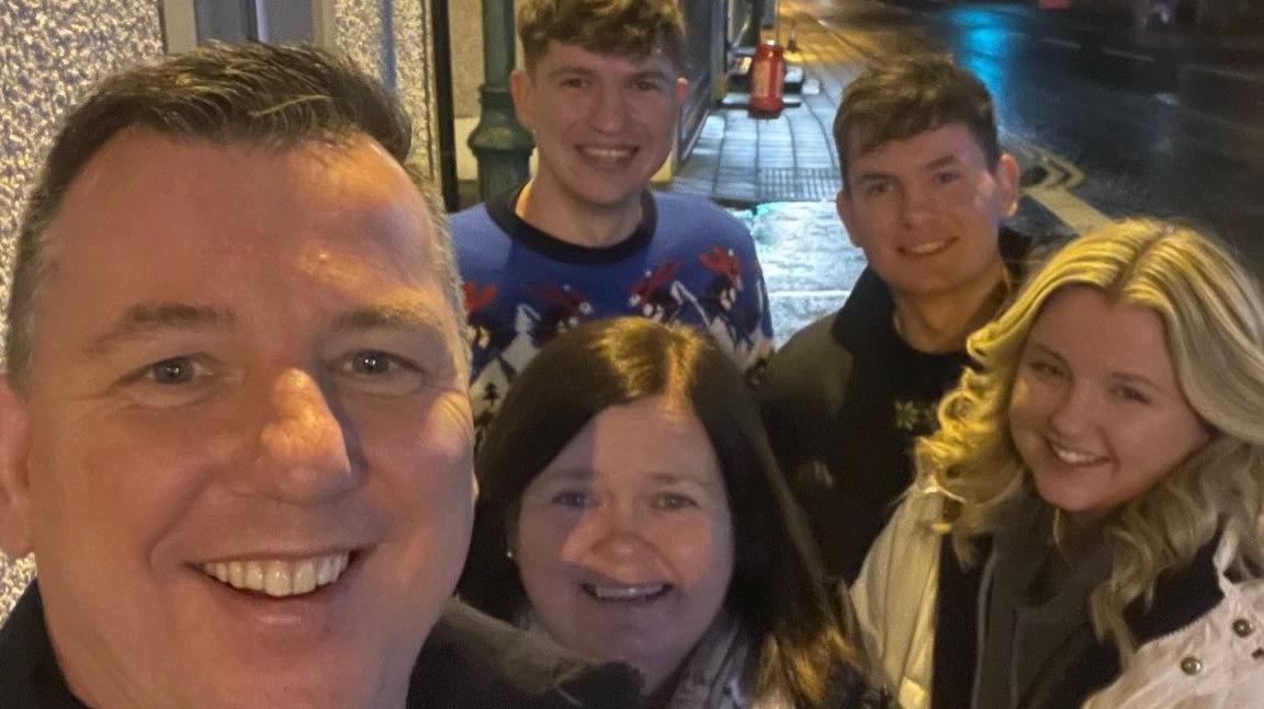 Glenn Johnson's selfie with his wife and children after they all helped to serve last year's Christmas Day dinner.  Glenn and his two sons all have short, dark hair and they are wearing Christmas jumpers. His wife and daughter have shoulder-length hair and they are wearing pale-coloured coats. 
 