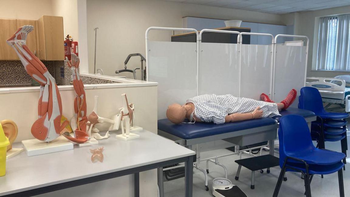 A replica hospital ward with a bed with a dummy lying on it, in front of a white screen. There are two blue chairs in the foreground and replica body parts to teach anatomy on a white table