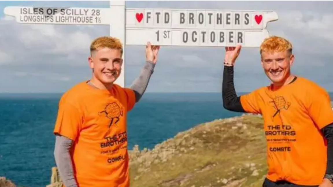 Two men in orange t-shirts standing in front of the sign for Land's End, on a cliff top overlooking the sea. The t-shirts are printed with a logo of a running brain, with The FTD Brothers written underneath. The Land's End sign has been customised to say "FTD Brothers, 1st October".