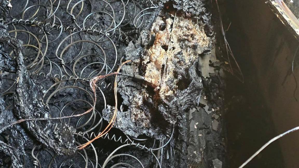 Close up of exposed metal springs of a burnt, blackened and charred mattress. On top, a damaged charging cable can be seen alongside the remains of a melted tablet.