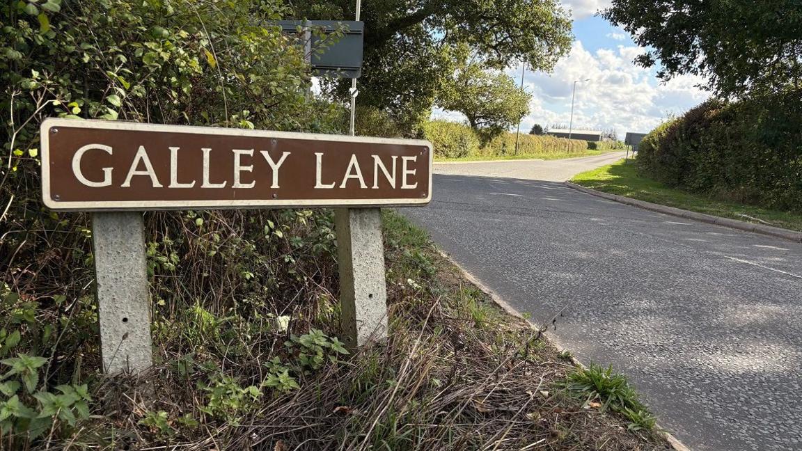 A brown sign in the foreground with GALLEY LANE in white letters is in the grass verge alongside a country road lined by hedges.
