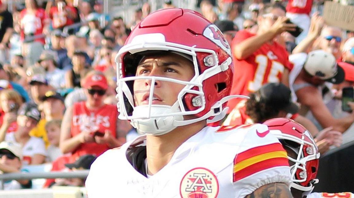 Louis Rees-Zammit enters the field in the red and white kit of the Kansas City Chiefs. A red number 9 is on his white shirt and he wears a red American football helmet. Louis wears shoulder pads and has a heavily tattooed right arm. A crowd dressed in red cheers behind him. 