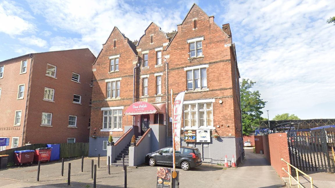 A red-brickhotel with stairs leading to the entrance,bins on the left, and a metal fence with trees on the right under a partly cloudy sky.