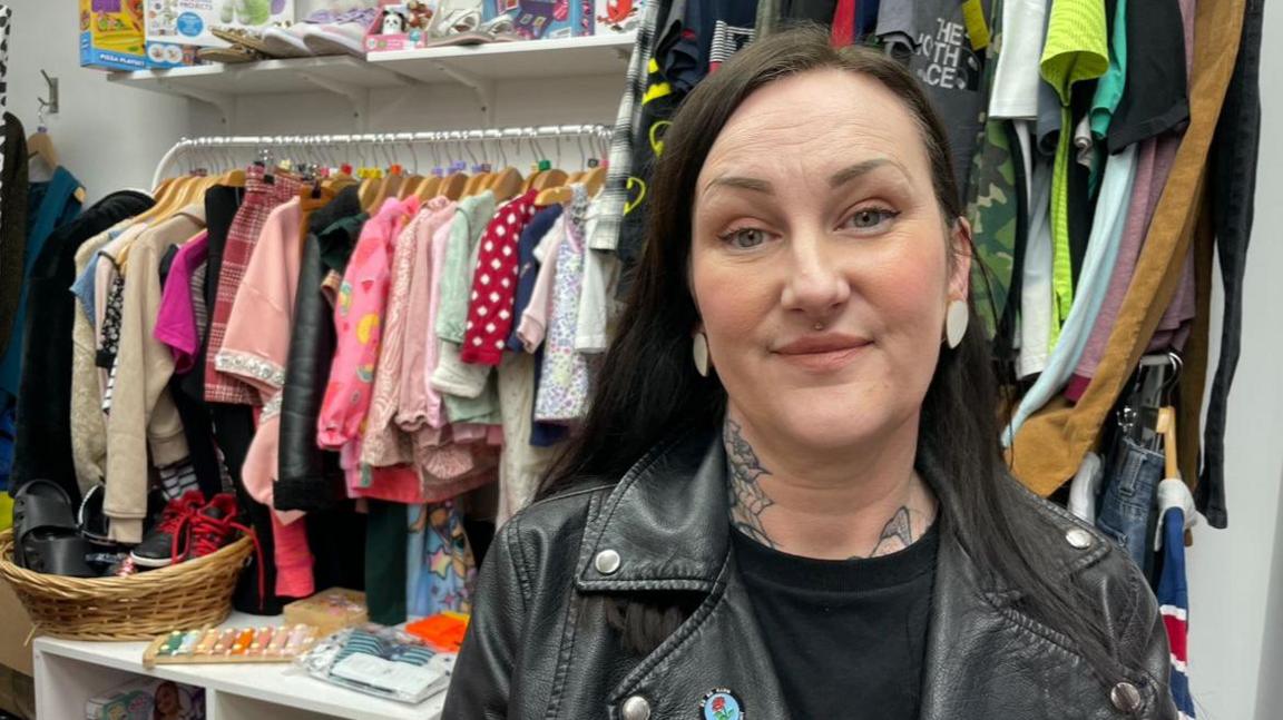 A woman with dark hair, wearing a leather jacket stands in front of a rail of secondhand children's clothes.