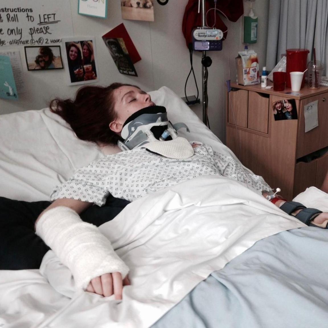 A woman with red hair lies on a hospital bed with a neck brace and a cast on her hand. There are photos stuck to the wall above her bed.