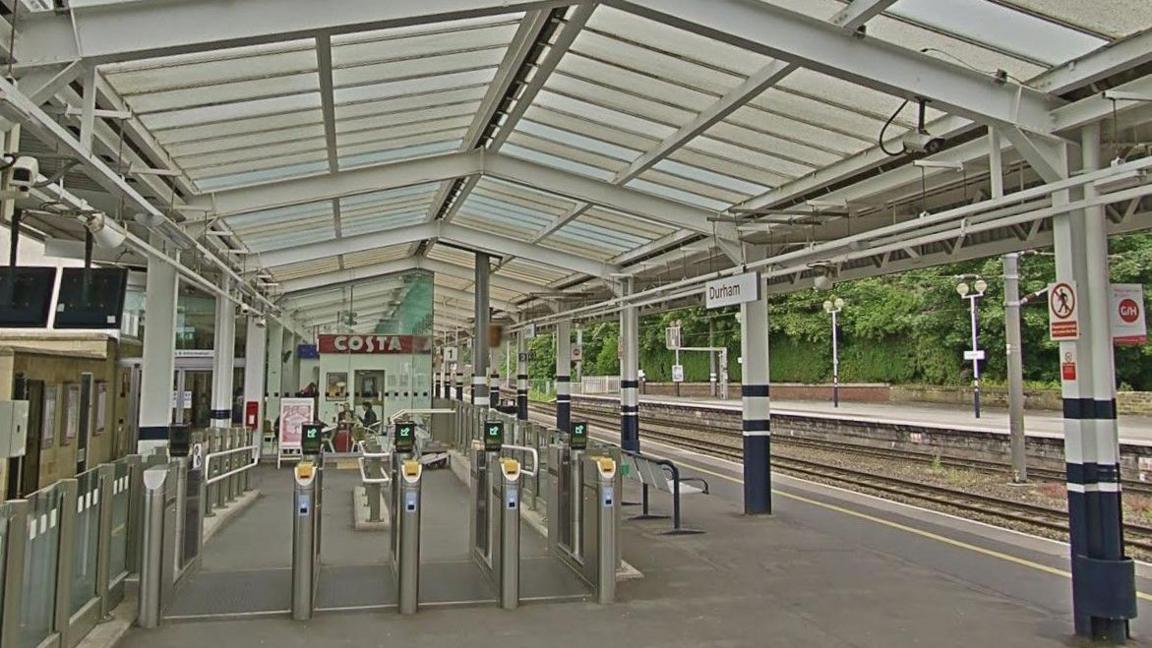 A platform at Durham station. It is covered by a large glass roof supported by metal poles with ticket barriers in the foreground.