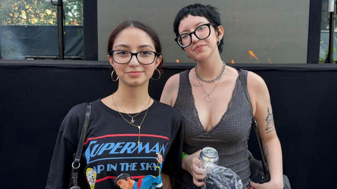 Two young women with glasses stand in front of a barrier, smiling. One wears a black t-shirt with a Superman graphic print on it, while the other wears a grey textured vest top. 