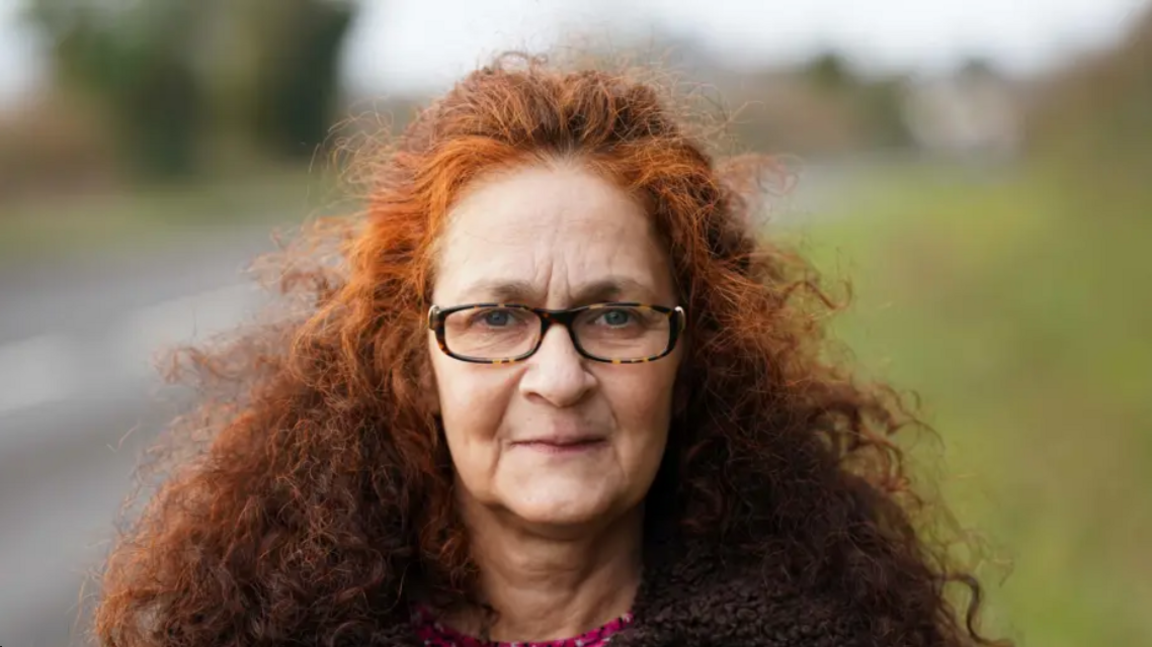 A woman with red curly long hair and is wearing glasses. she is wearing a dark brown teddy jacket and has a pink floral pattern shirt underneath it.