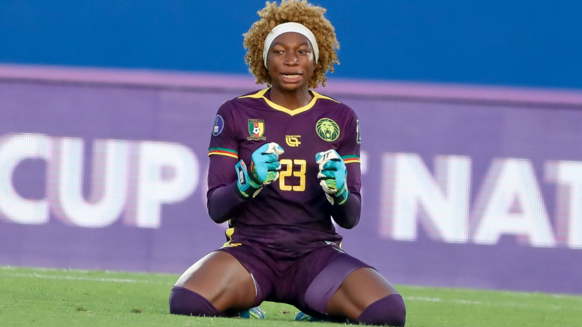 Michaely Bihina kneels back on her heels on a football pitch as she clasps her two hands in front of her body in celebration during a match at Wafcon 2026. The goalkeeper wears a white hairband, an all-purple kit, including undersleeves, and blue gloves