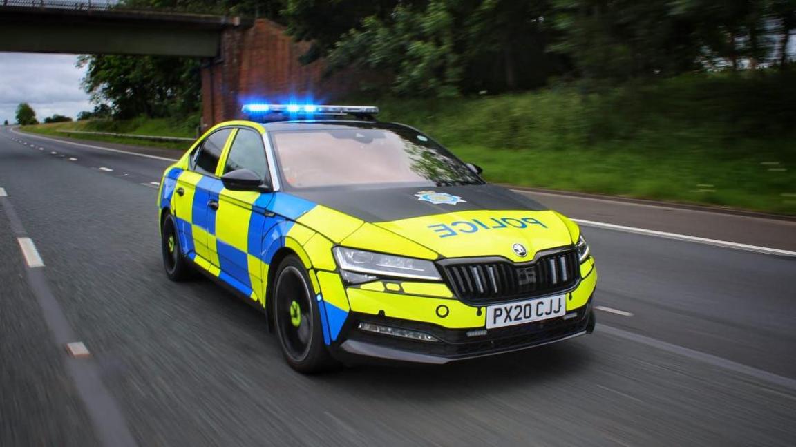 A Cumbria Police car being driven along a motorway. The vehicle has a yellow and blue livery and the blue lights on its roof are flashing. A bridge over the road is in the background and trees and grass are to the side of the road.