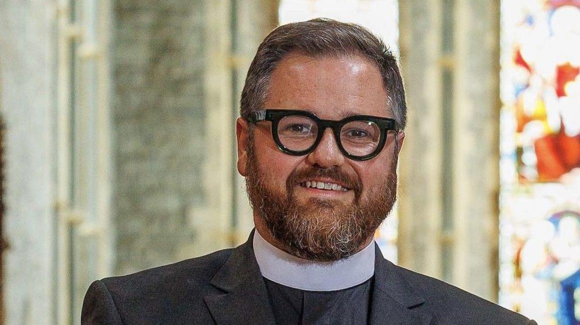 A man in a suit and white collar smiling at the camera. He has a beard and is wearing thick-framed black glasses. In the background, a stained glass window is visible