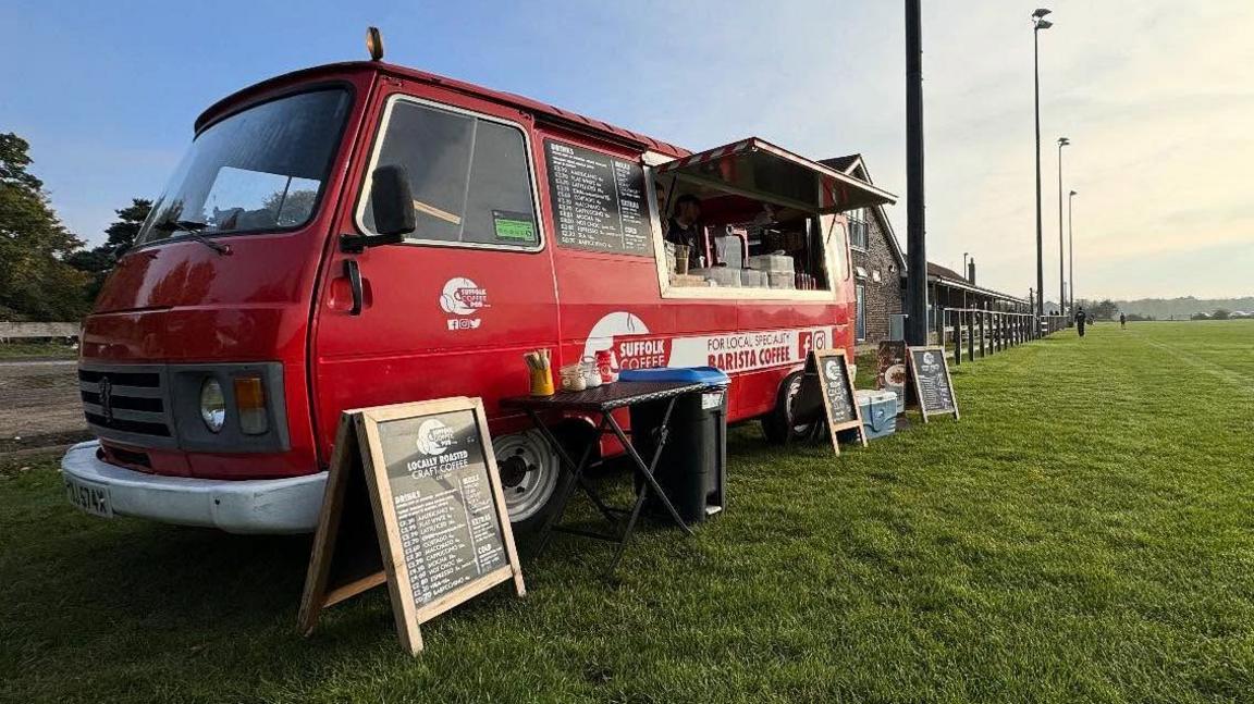 A red van was stood next to a wooden fence. On the side of the van was a window with signs on the side.