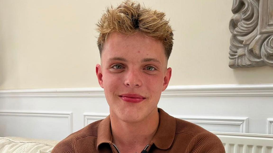 Samuel Goodman, a 20 year old, white man sitting on a sofa. Samuel is blonde and is wearing a brown knitted polo shirt.