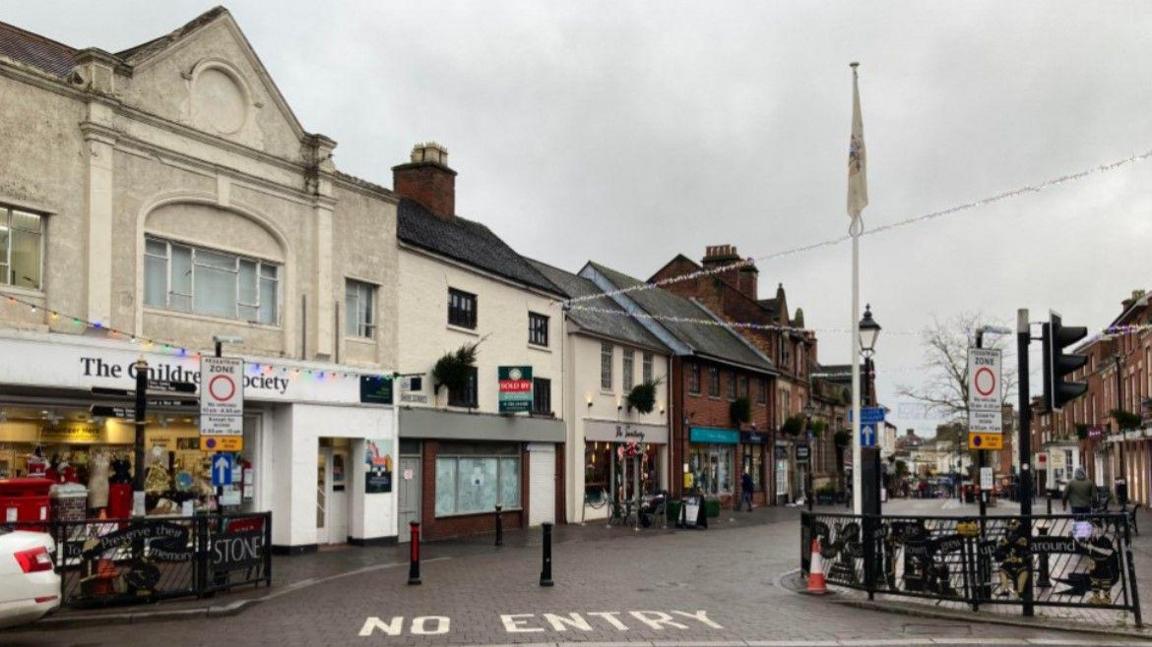 The entrance to Stone High Street, with two bollards, as well as the words no entry on the ground. Shops are prominent on the left.