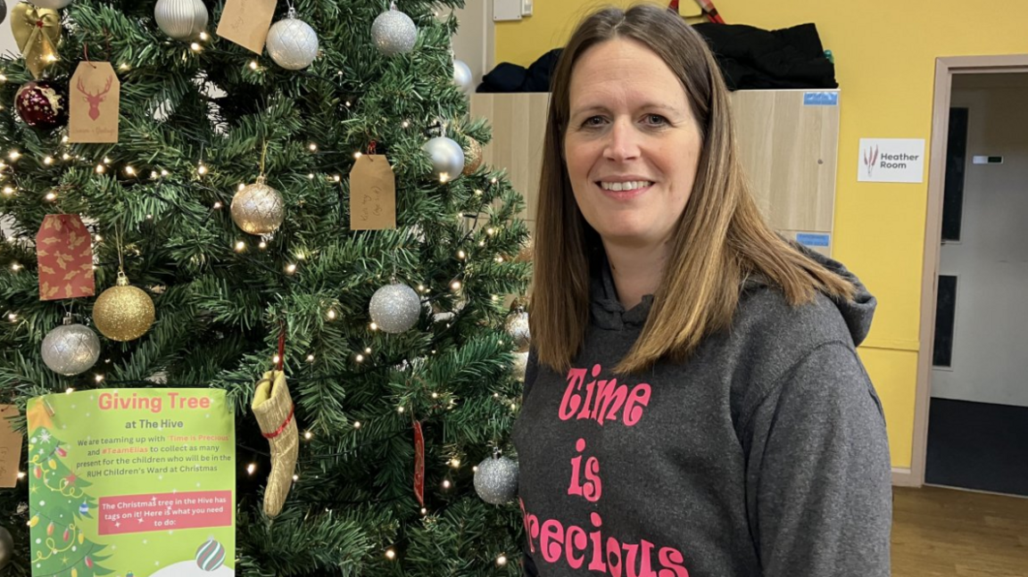 Nicky is pictured wearing a grey hoody with pink Time is Precious writing with a Christmas tree in the background. It has silver and gold baubles and little woollen stockings hanging on the branches.