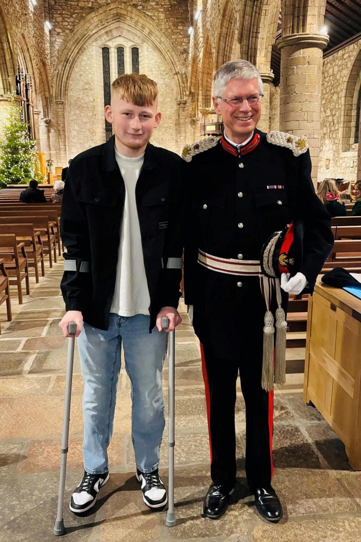 Teenage boy with ginger hair wearing light blue jeans, a light T-shirt and black casual jacket and trainers, standing on crutches, next to an older man with grey hair, smiling, wearing a dark Lord Lieutenant uniform, both standing in the aisle of a cathedral.