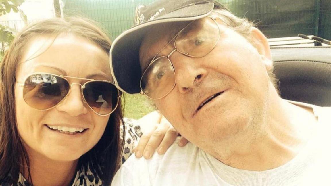 Dr Becs Bradford with her father, Richard. They are pictured in a selfie, with their faces close to the camera. Becs has long dark hair and is wearing sunglasses. Her hand is on her father's shoulder. Richard is leaning his head in close to hers and smiling. He is wearing a cap and glasses. 